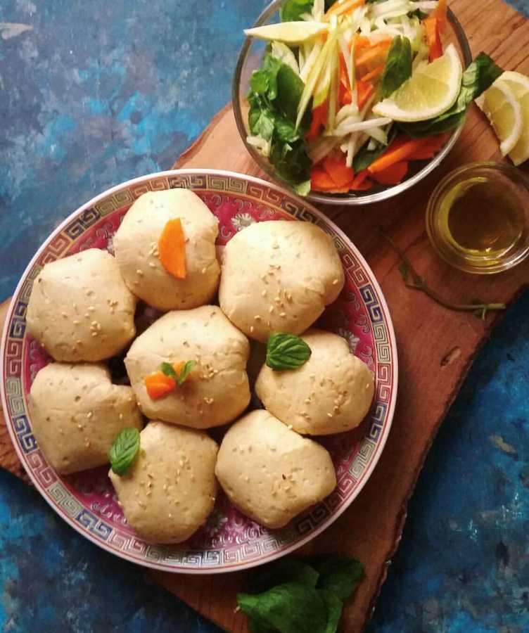 A plate of round, sesame-topped buns sits next to a bowl of salad and a small dish of oil on a wooden board.