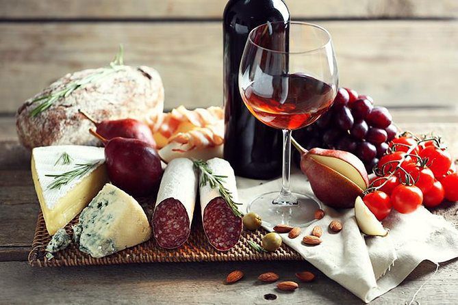 A wooden board holds wine, cheese, salami, bread, grapes, tomatoes, pears, and almonds on a rustic surface.