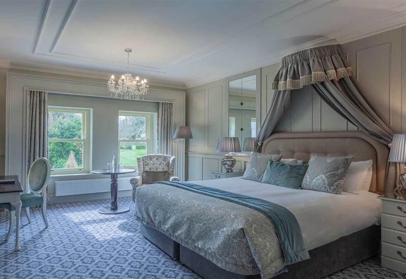 A bedroom features a bed with a canopy, a patterned carpet, a small table with two chairs, and two windows overlooking trees.