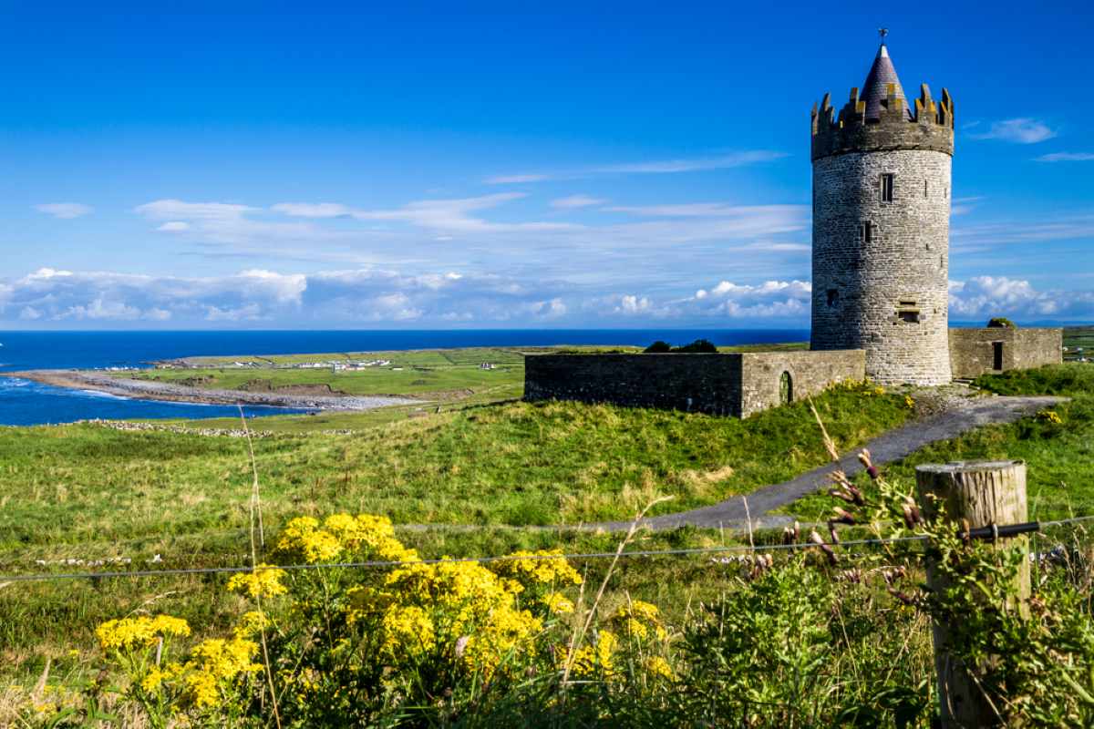 A stone tower stands on a grassy hill overlooking the ocean under a bright blue sky.