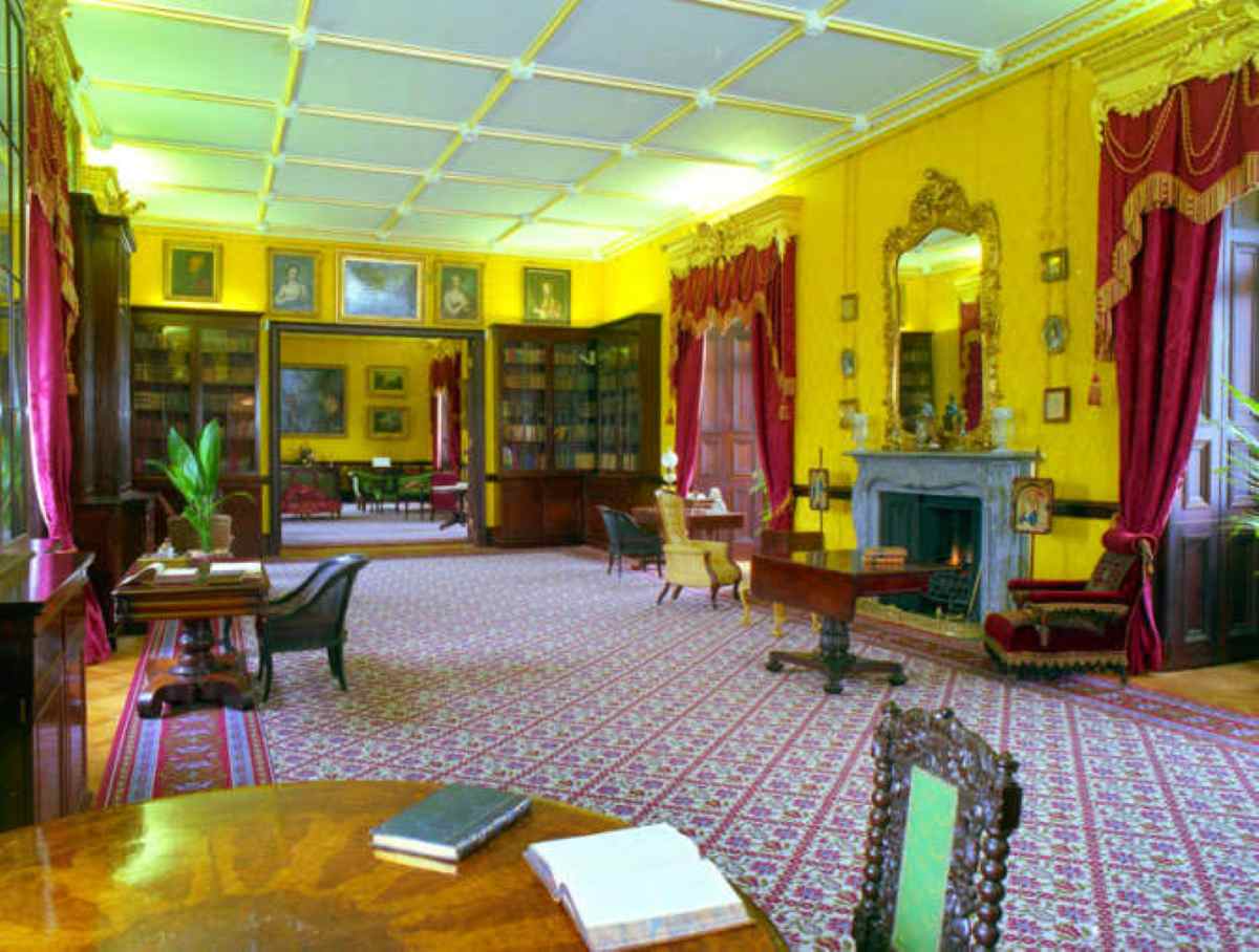 A spacious room with yellow walls, red curtains, a fireplace, and bookshelves features patterned carpet and antique