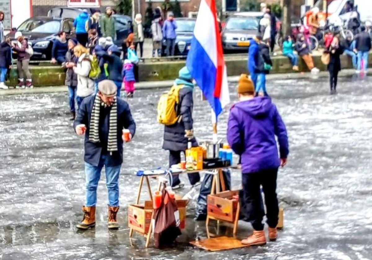 People stand on a frozen canal surface near a small table with drinks and a Dutch flag.