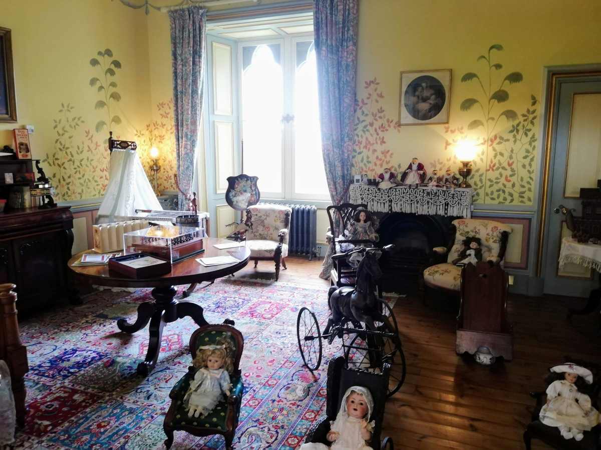 A room with yellow floral wallpaper, a central table, a fireplace, and several antique dolls and a rocking horse.