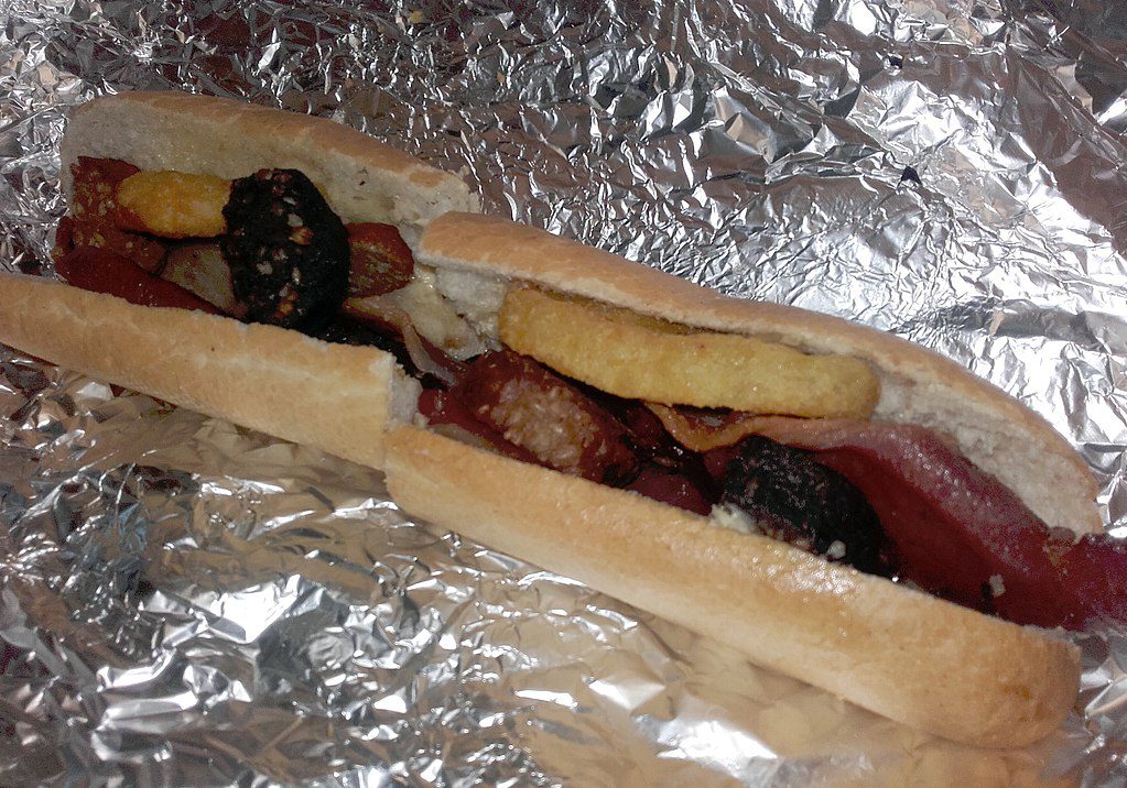 A long bread roll filled with sausage, bacon, black pudding, and a hash brown rests on a sheet of aluminum foil.