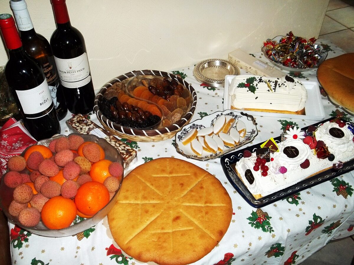 A table is set with wine bottles, a bowl of fruit, dried fruits, pastries, and two decorated cream cakes.