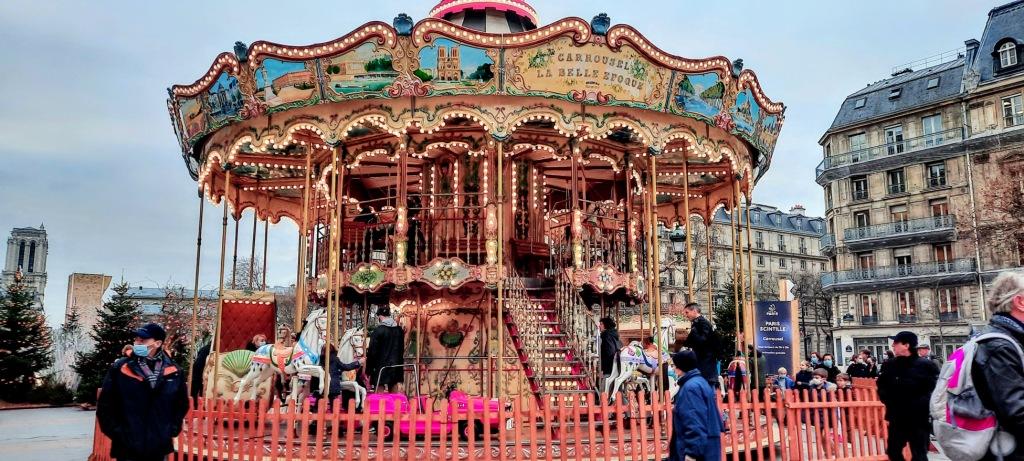 An ornate, multi-level carousel with horse figures stands in a city square surrounded by people and historic buildings.