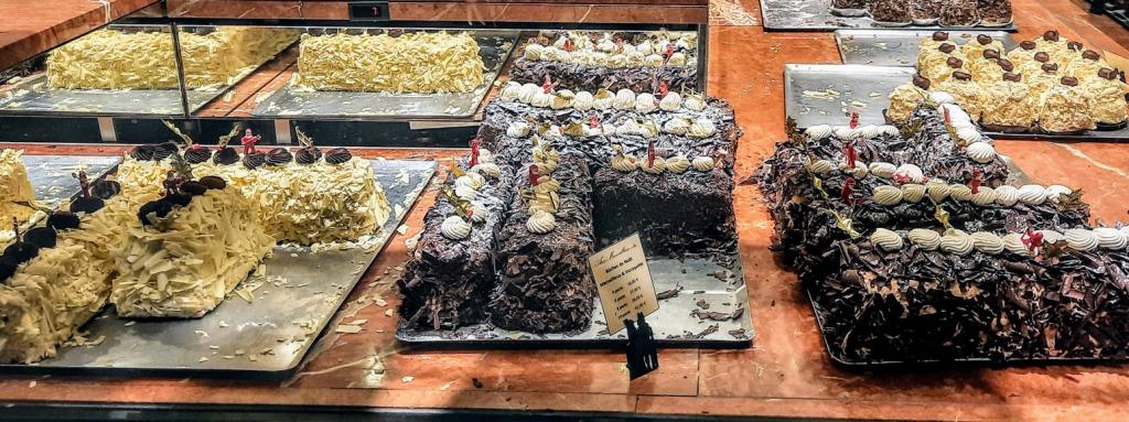 Trays of various chocolate and white chocolate log cakes are displayed on a counter with small decorative figurines.
