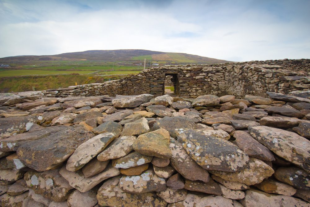 A dry stone wall with a small doorway stands in front of a grassy hillside under a cloudy sky.