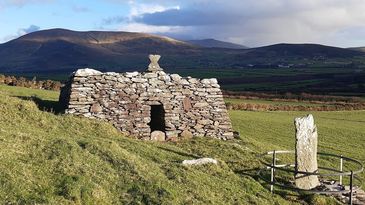 A stone structure with a small doorway sits in a grassy field near a fenced standing stone, with mountains in the distance.
