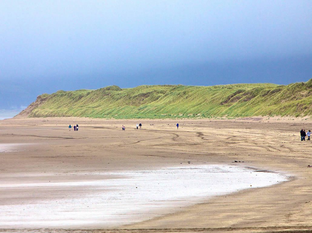 People walk along a wide, sandy beach beneath a grassy dune under a cloudy sky.