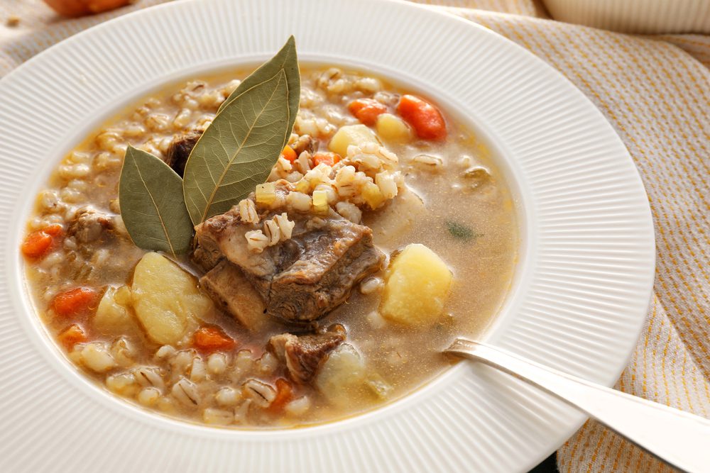 A bowl of soup containing meat, potatoes, carrots, and grains, garnished with two bay leaves.