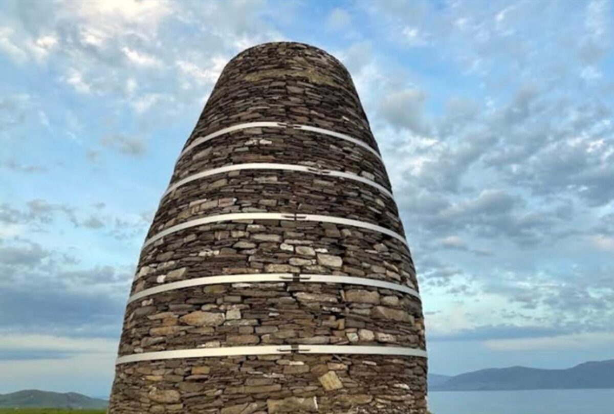 A conical stone structure reinforced with horizontal metal bands stands against a cloudy sky and a distant body of water.