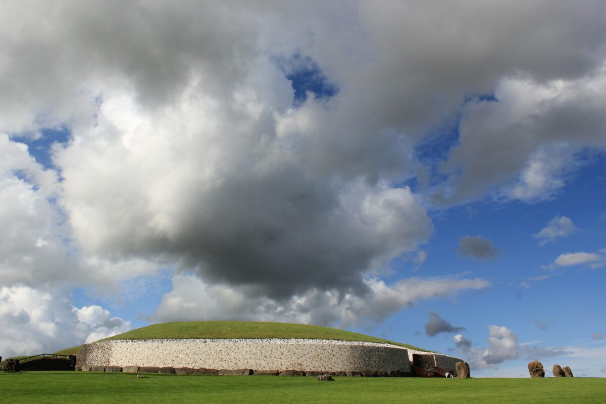 A grassy mound with a stone-walled base sits under a large, dramatic cloud formation in a bright blue sky.