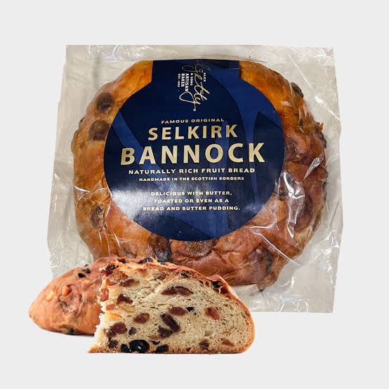 A round loaf of fruit bread in plastic packaging sits behind a slice of the same bread on a white background.
