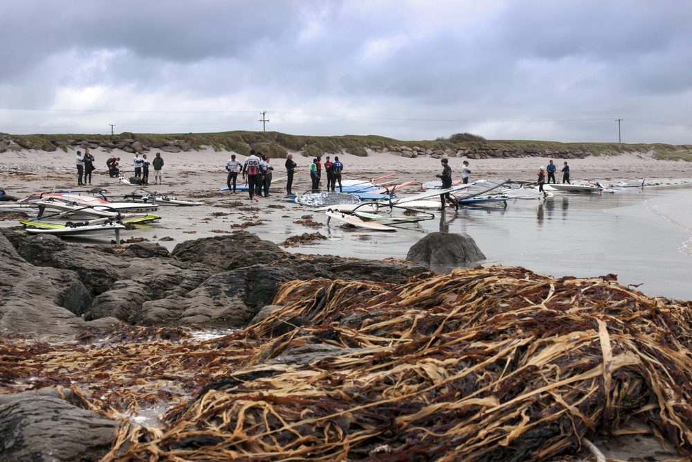 People in wetsuits gather on a sandy beach with windsurfing equipment near rocky, seaweed-covered terrain.