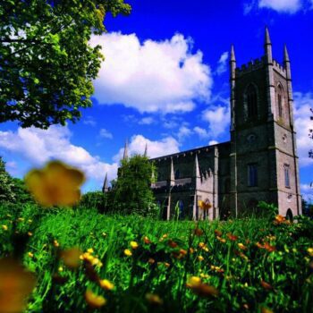 Down Cathedral in Downpatrick Northern Ireland. A square boxy tower at the front with castellation around the top and each side has a pointed steeple. There is a garden of yellow buttercups in the foreground and the sky is bright blue