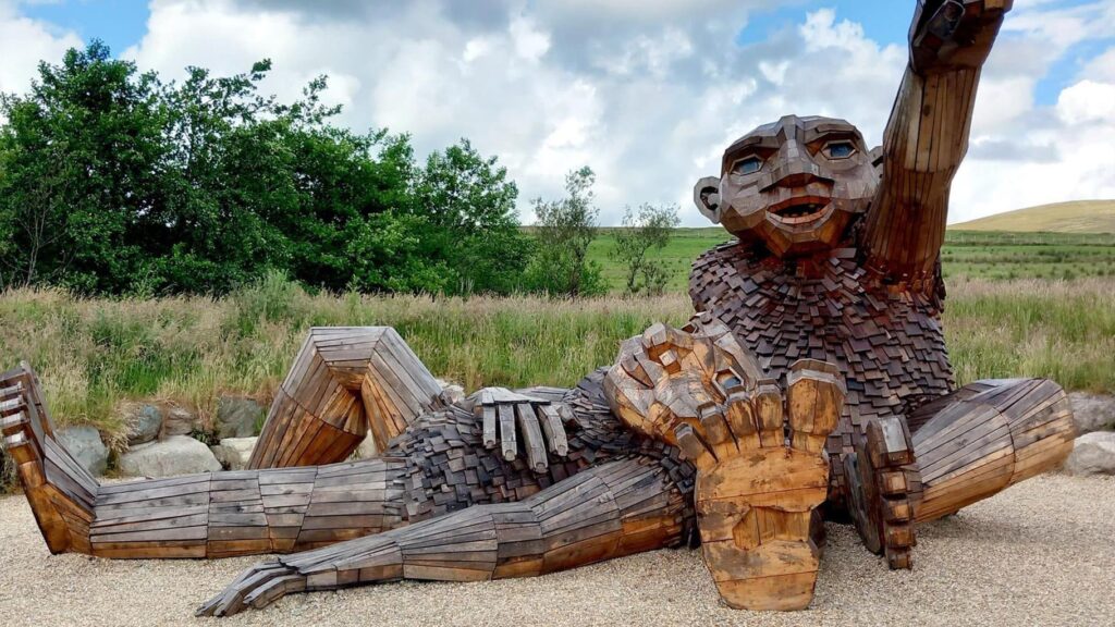 A wooden carving on the Sperrins sculpture trail. the wooden troll is lying down and waving with a green grass field behind him