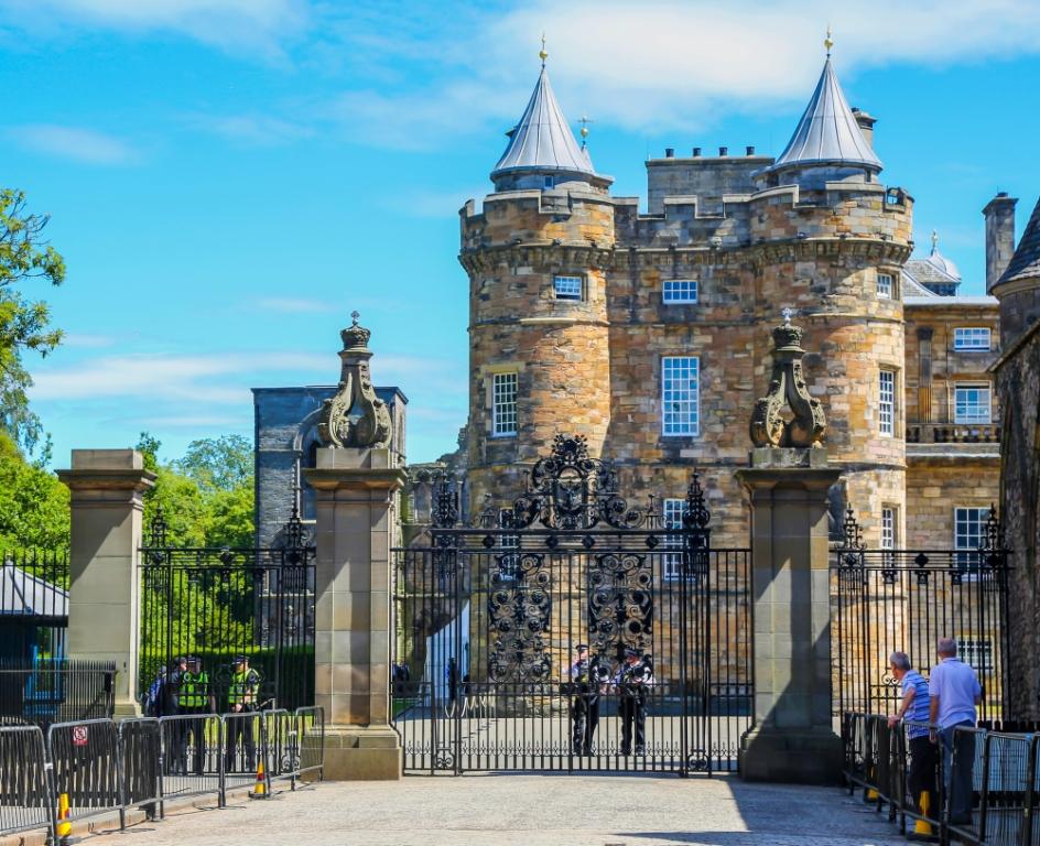The grand palace of Holyrood. Guards stand behind an ornate wrought iron gate. The palace itself has two round towers with peaked roofs