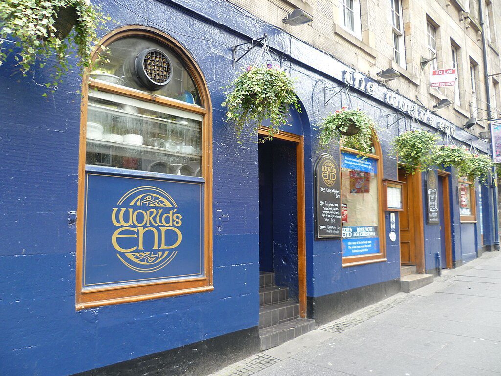 A blue painted pub with gold lettering on the arched windows and green hanging baskets across the front of the building