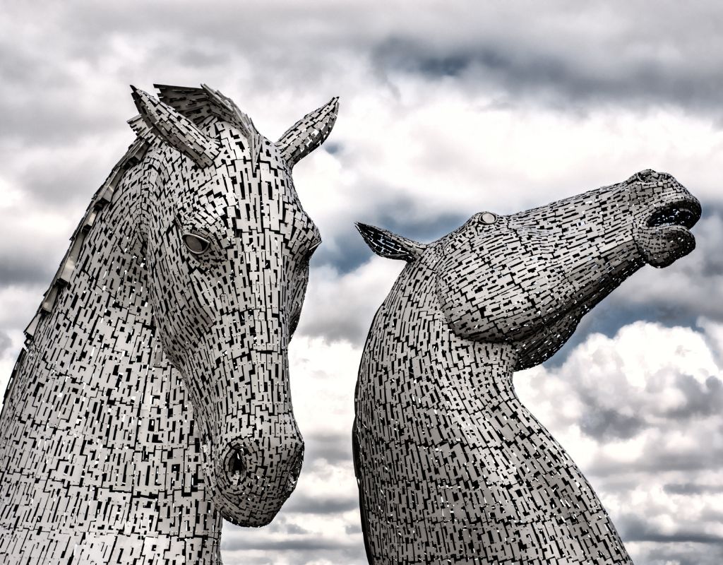 The Kelpies are towering, 30-meter-high sculptures depicting the heads of two horses, intricately designed with sleek, modern lines and a stainless steel finish that reflects light, giving them a dynamic and powerful appearance as they rise dramatically above the landscape.