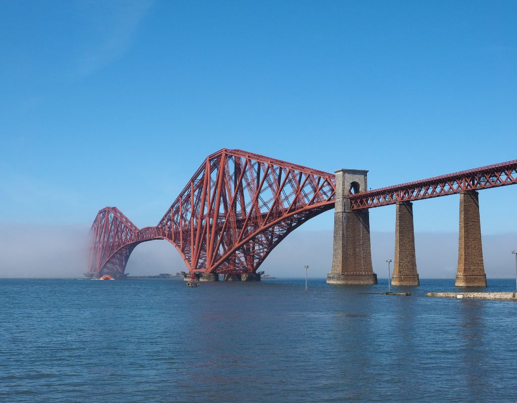 The Firth of Forth Bridge, located in Scotland, is an iconic cantilever railway bridge characterized by its distinctive red color and massive steel structure, featuring three towering double cantilevers connected by two central girders, spanning the Firth of Forth estuary with a striking and industrial elegance.