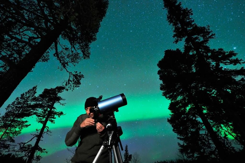 A photographer is taking pictures of the Northern lights in Ireland. The sky behind him is running with bright green and purple lights and the trees are silhouetted in black
