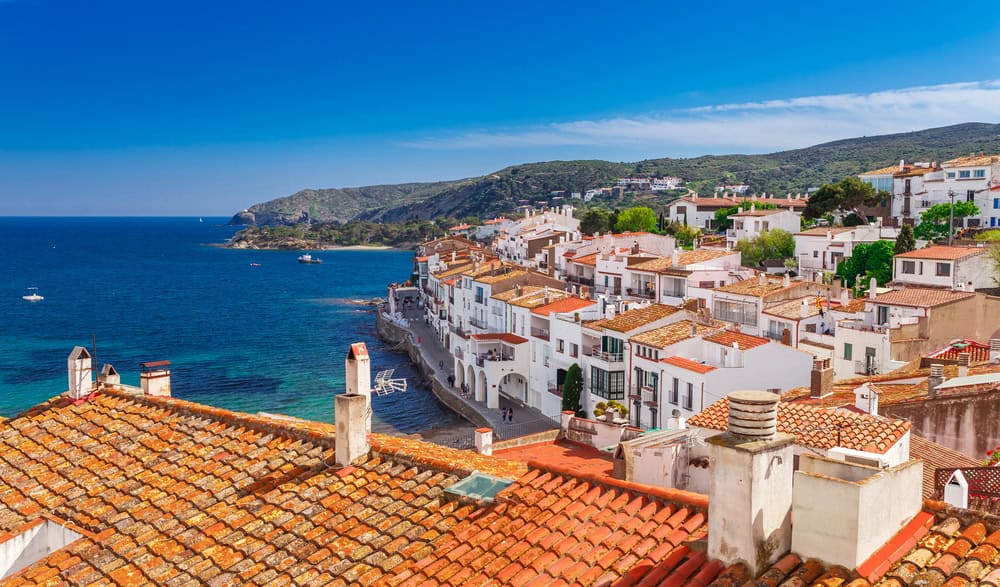 A view of Cadaques from above with terracotta roof tiles, hitewashed houses the rolling hills in the background and a deep blue green bay in front of the houses. Top view in Cadaques, Catalonia, Spain near of Barcelona. Scenic old town with nice beach and clear blue water in bay. Famous tourist destination in Costa Brava with Salvador Dali landmark