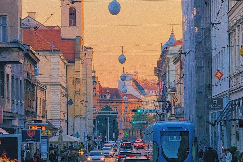 A vibrant urban street scene in Zagreb featuring trams, cars, and pedestrians during twilight.