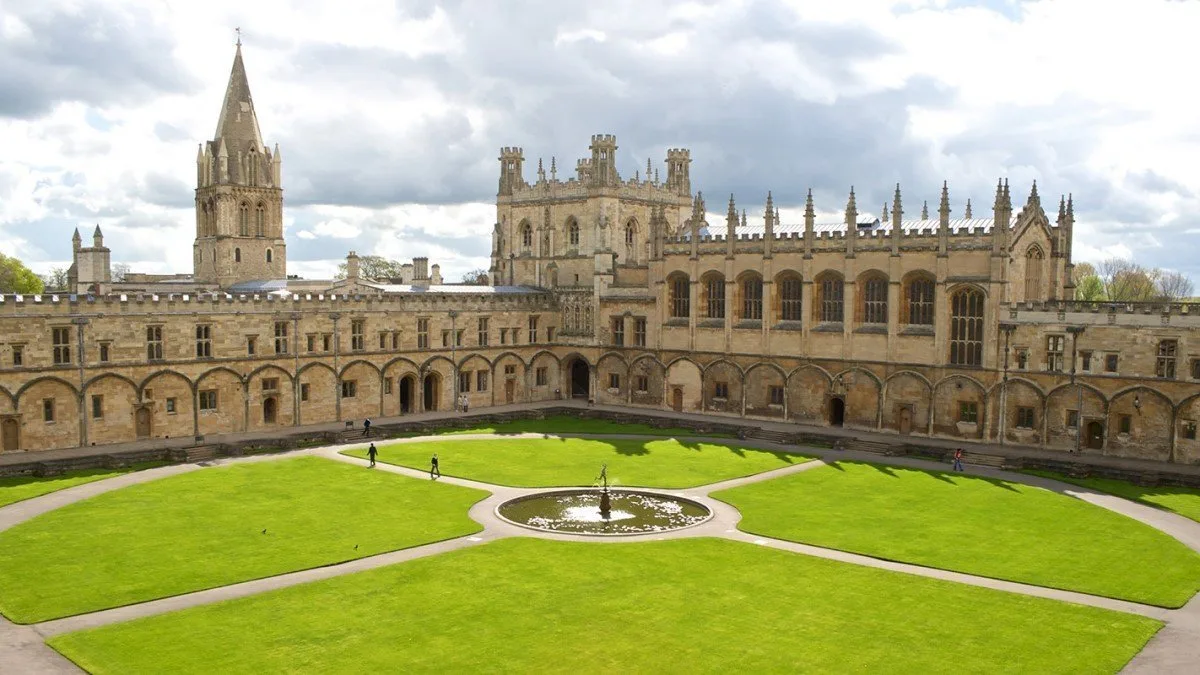 A large stone building with a central courtyard, a circular fountain, and a grassy lawn under a cloudy sky.