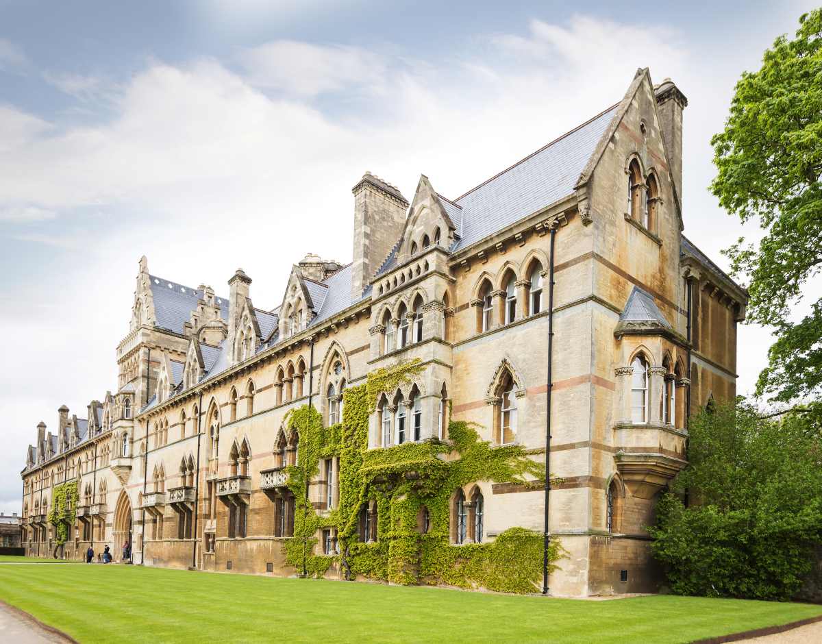 A large, multi-story stone building with Gothic architectural features and ivy climbing its walls sits beside a green lawn.
