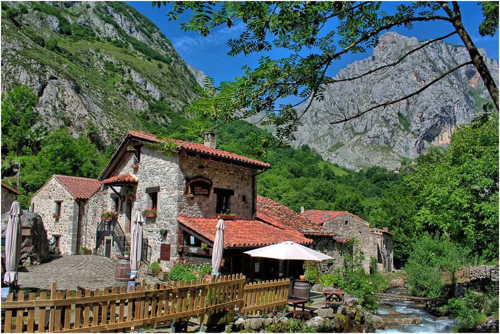 Tazones, Asturia a terracotta tiled roof house built of stone with a patio with tables chairs umbrellas and poted plants sits in front of a craggy mountin