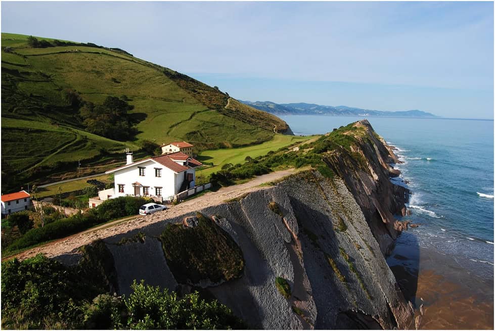 A house sits perched on a granite cliff with a deep green mountain behind it. The house has great views of the deep blue sea