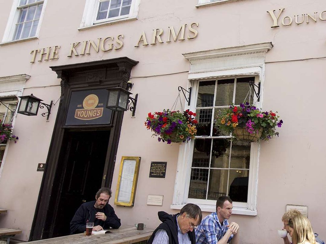 People sit at outdoor tables in front of a pale pink building labeled The Kings Arms with hanging flower baskets.