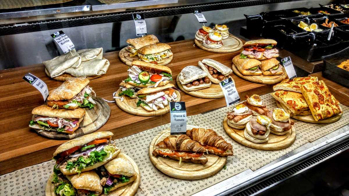 A vibrant deli counter in the UK or Ireland showcases fresh sandwiches, wraps, focaccia, and breakfast muffins.
