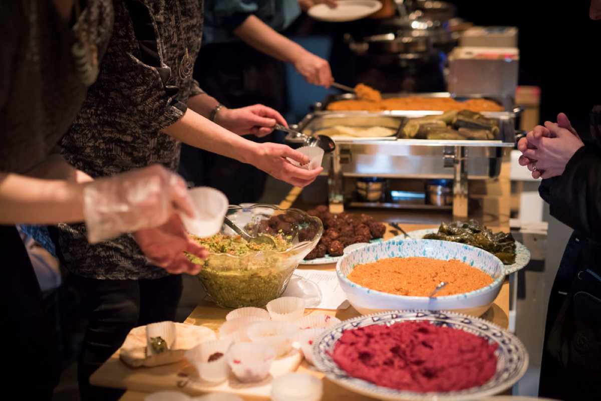People serve themselves food from a buffet table featuring various bowls of dips, salads, and warm dishes.