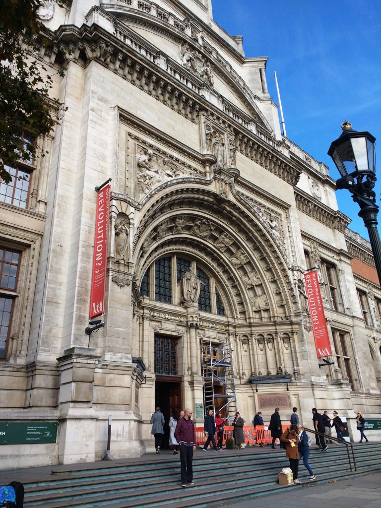 Front entrance of V&A museum in things to do in Chelsea. The building is white stone with red banners hanging rom its frontage