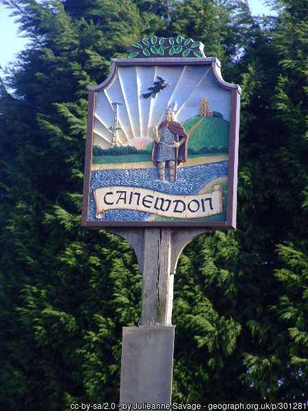 A wooden village sign for Canewdon features a carved figure in a horned helmet standing before a sunburst and a tower.