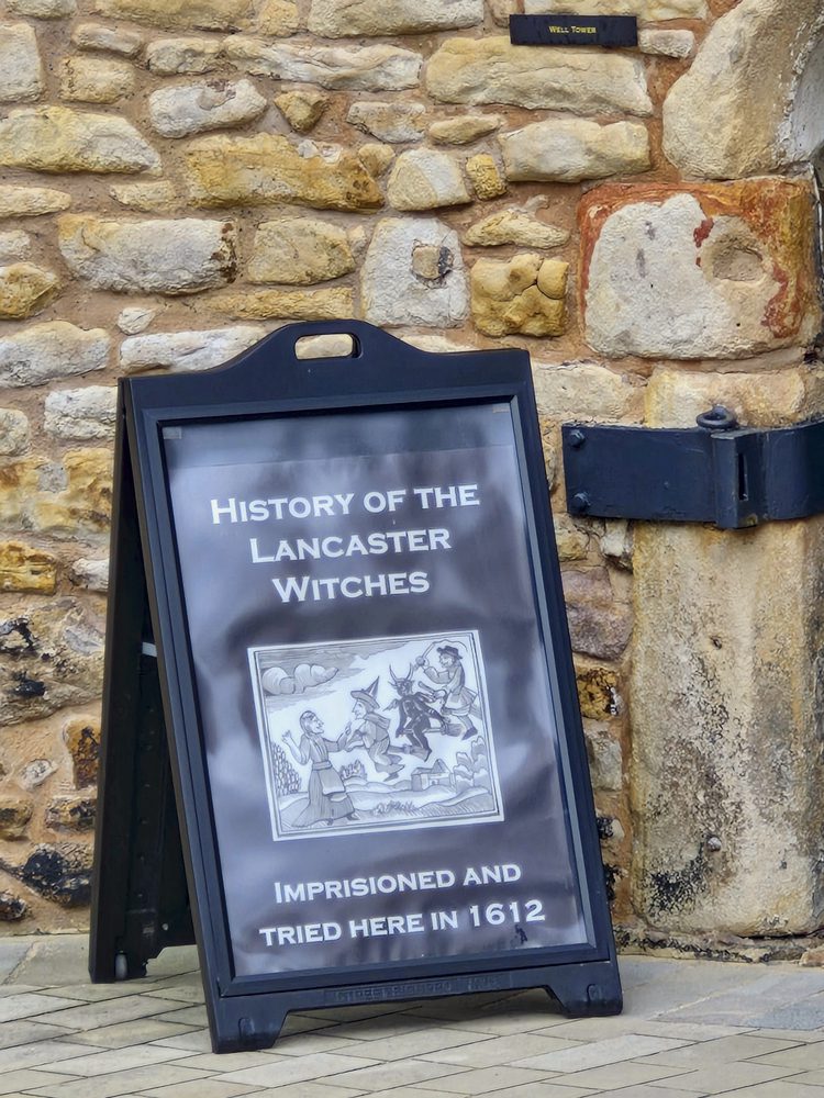 A black A-frame sign against a stone wall reads History of the Lancaster Witches, imprisoned and tried in 1612.