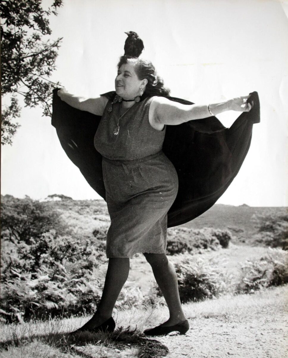 A woman in a dress and cape walks outdoors with a bird perched on her head.