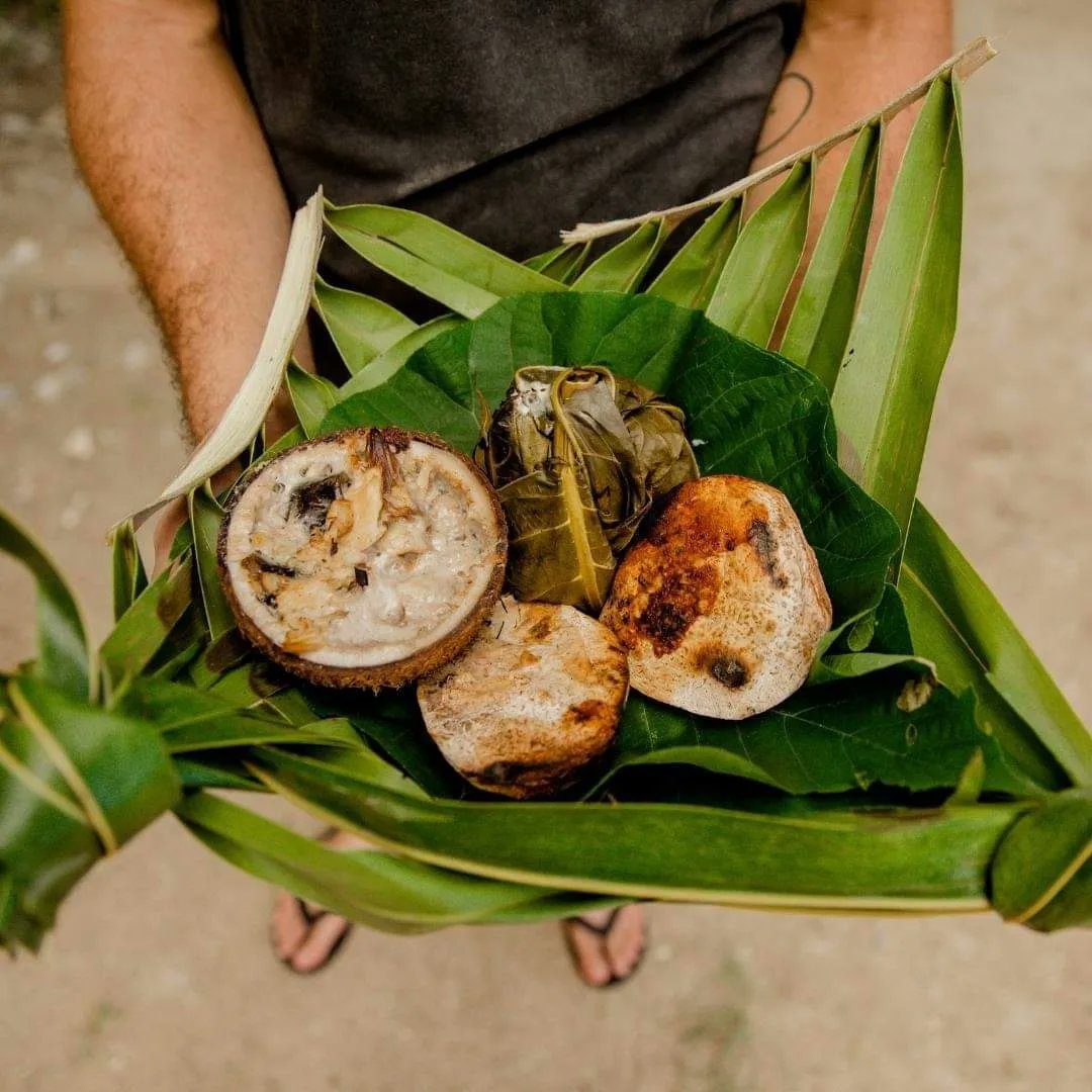 A person holds a woven palm leaf basket filled with traditional tropical foods, including a halved coconut and roasted items.