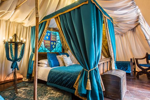 A bed with teal curtains and a heraldic crest sits inside a tent-like structure with draped fabric walls.