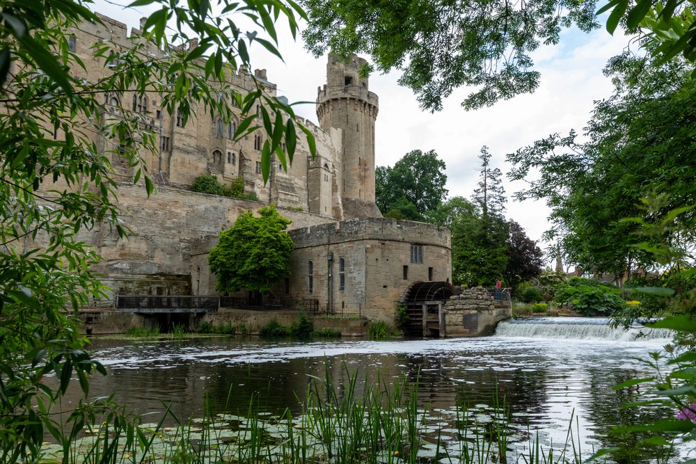 A stone castle with a tall tower overlooks a river with a small waterfall and a water wheel.