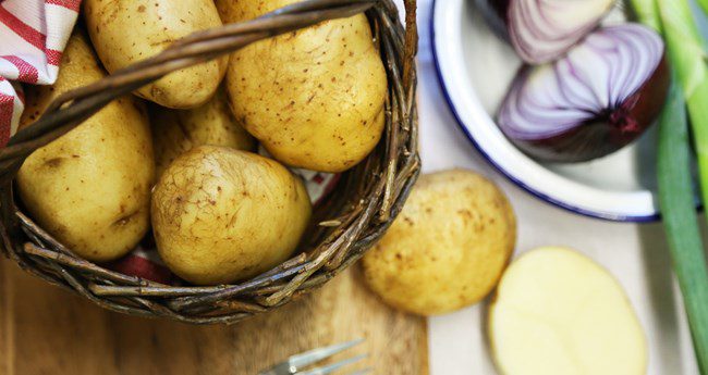 A wicker basket filled with raw potatoes sits on a wooden surface next to red onion wedges and green onions. Traditional Irish food