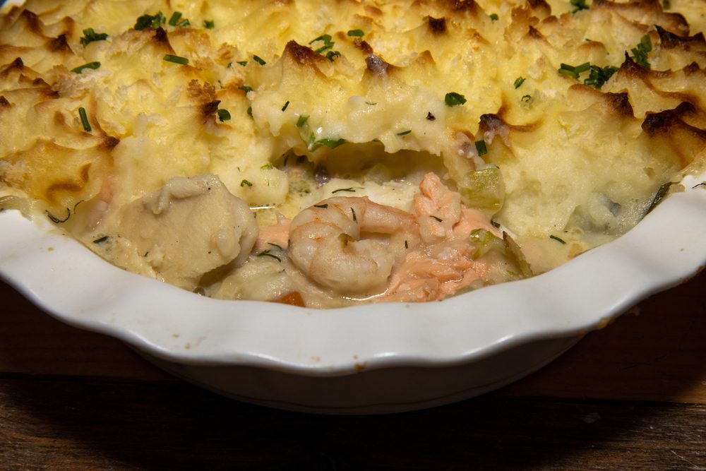 A seafood pie with a mashed potato topping sits in a white scalloped baking dish.