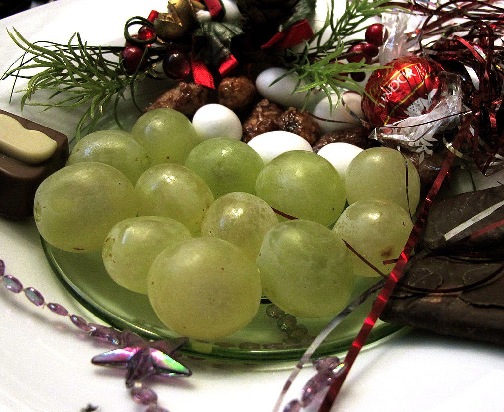 A glass plate holds green grapes, assorted chocolates, and nuts, decorated with festive greenery and red ribbons.