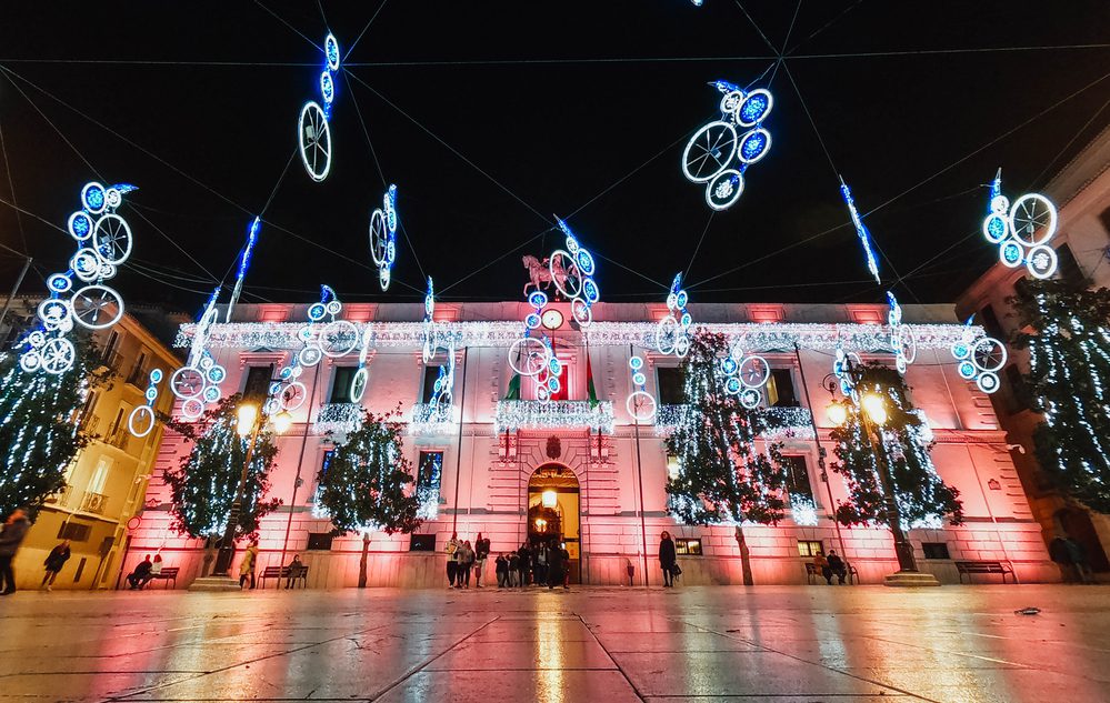 A building facade is illuminated with pink lights and decorated with hanging blue bicycle-shaped light displays at night. Granada, Spain - December The Town Hall of Granada in the Square of Carmen, Andalusia. Decorated street with Christmas lights