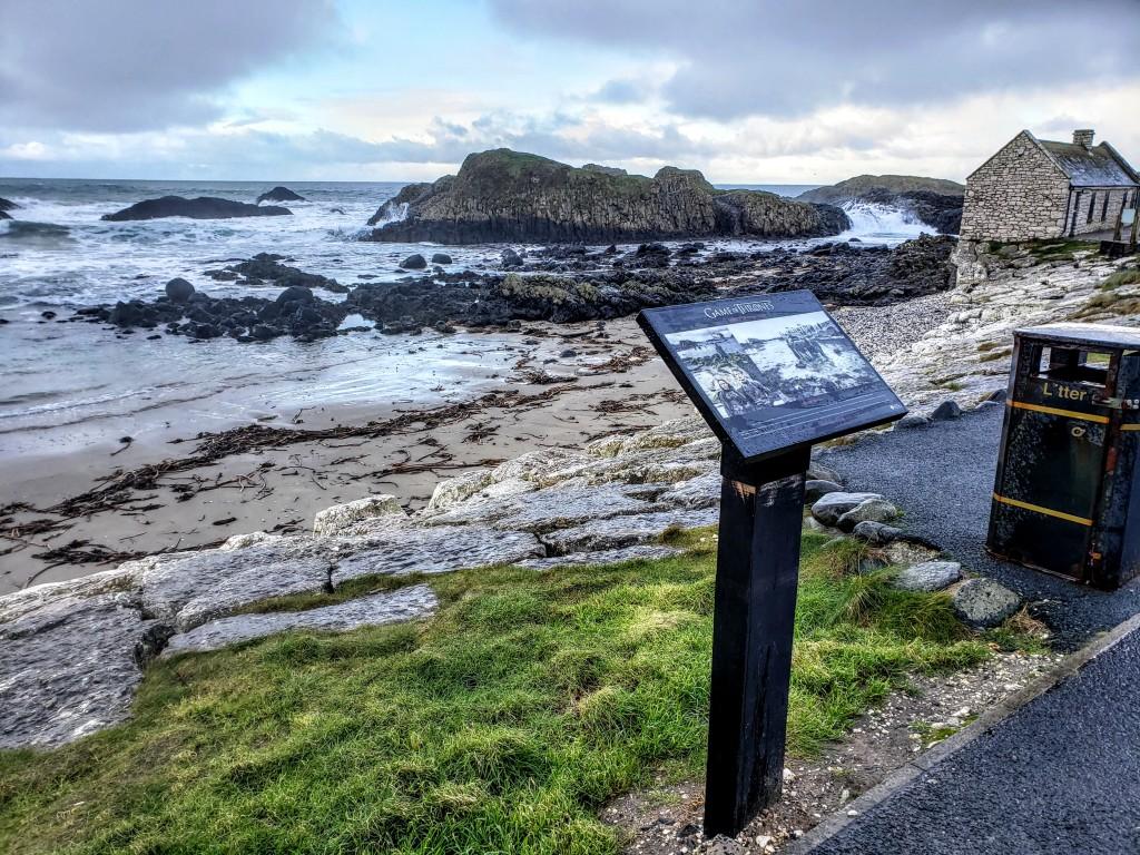 Coastal scene with rocky beach, informational sign, and stone building under cloudy sky.