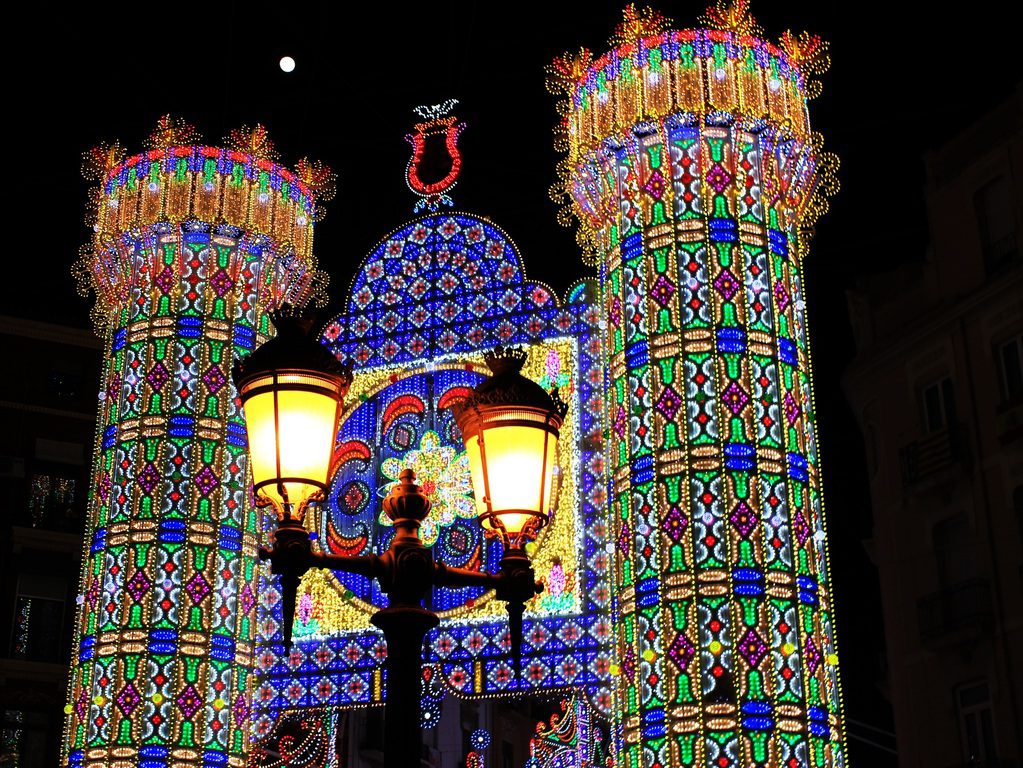 A church with two glowing lanterns stands before a large, ornate archway illuminated by colorful LED patterns at night.