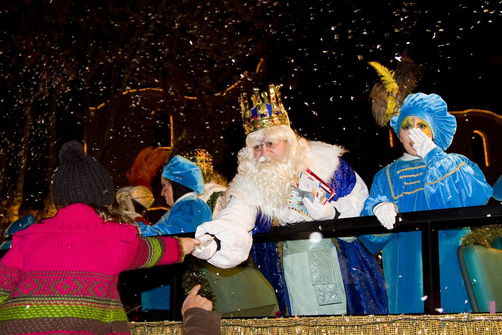 A person in a crown and white beard hands a letter to a child from a parade float as confetti falls at night.