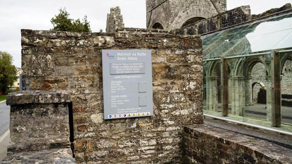 Stone wall with a sign for Boyle Abbey and a glass-covered ruin.